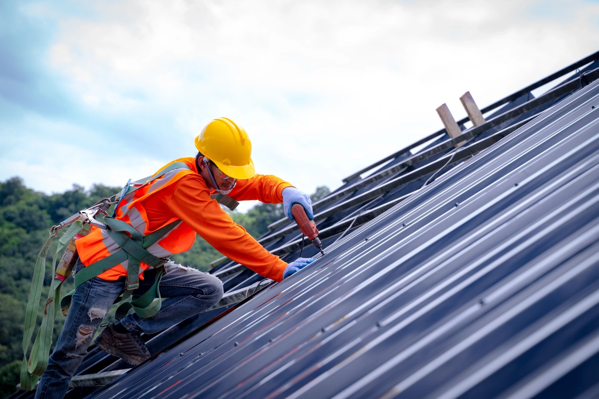Worker in orange vest and hard hat drills into metal roof panel wearing a safety harness
